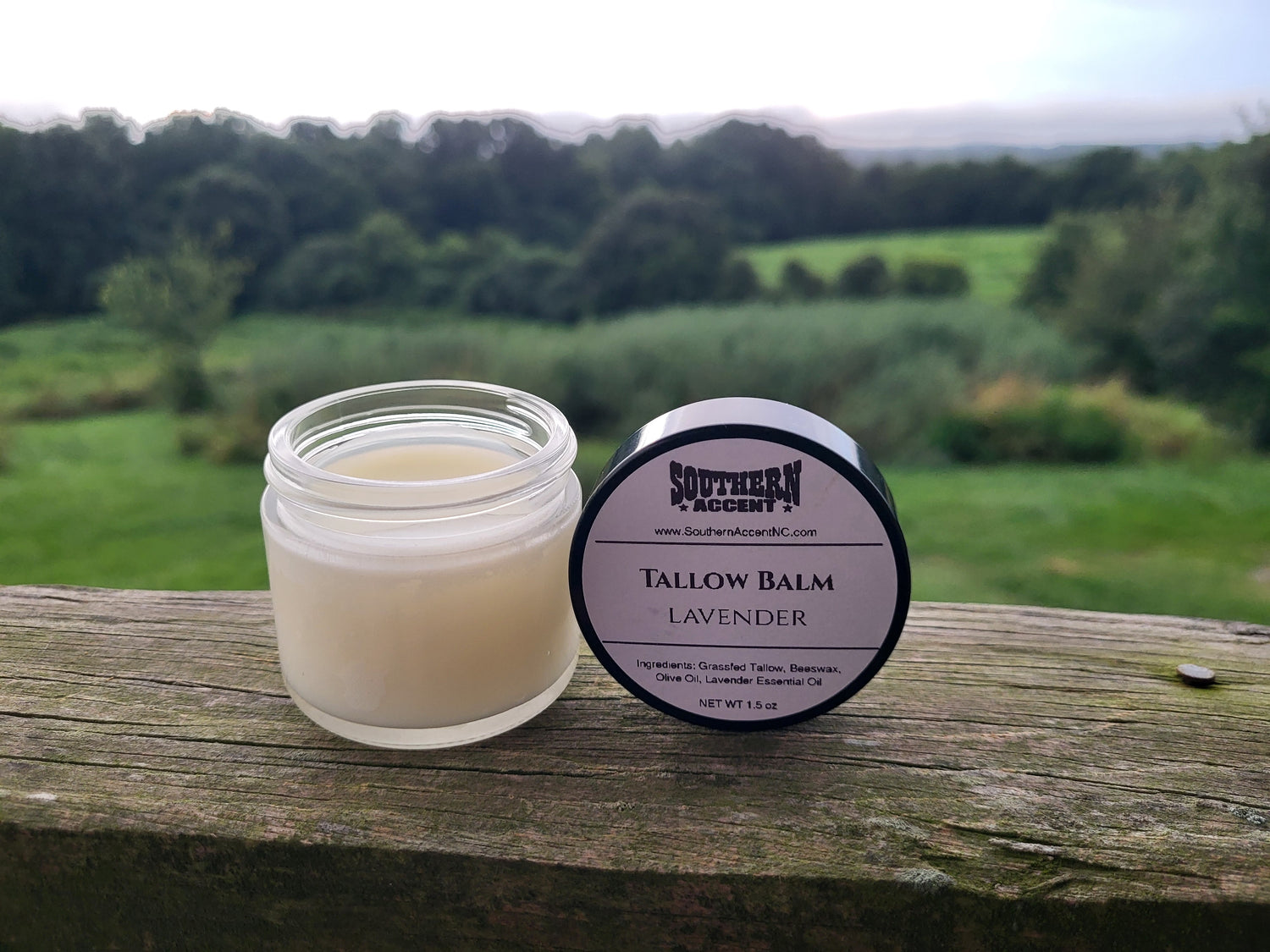 Clear Southern Accent Tallow balm jar showcasing the product inside and it's branded black lid on a rustic wooden surface with a scenic background