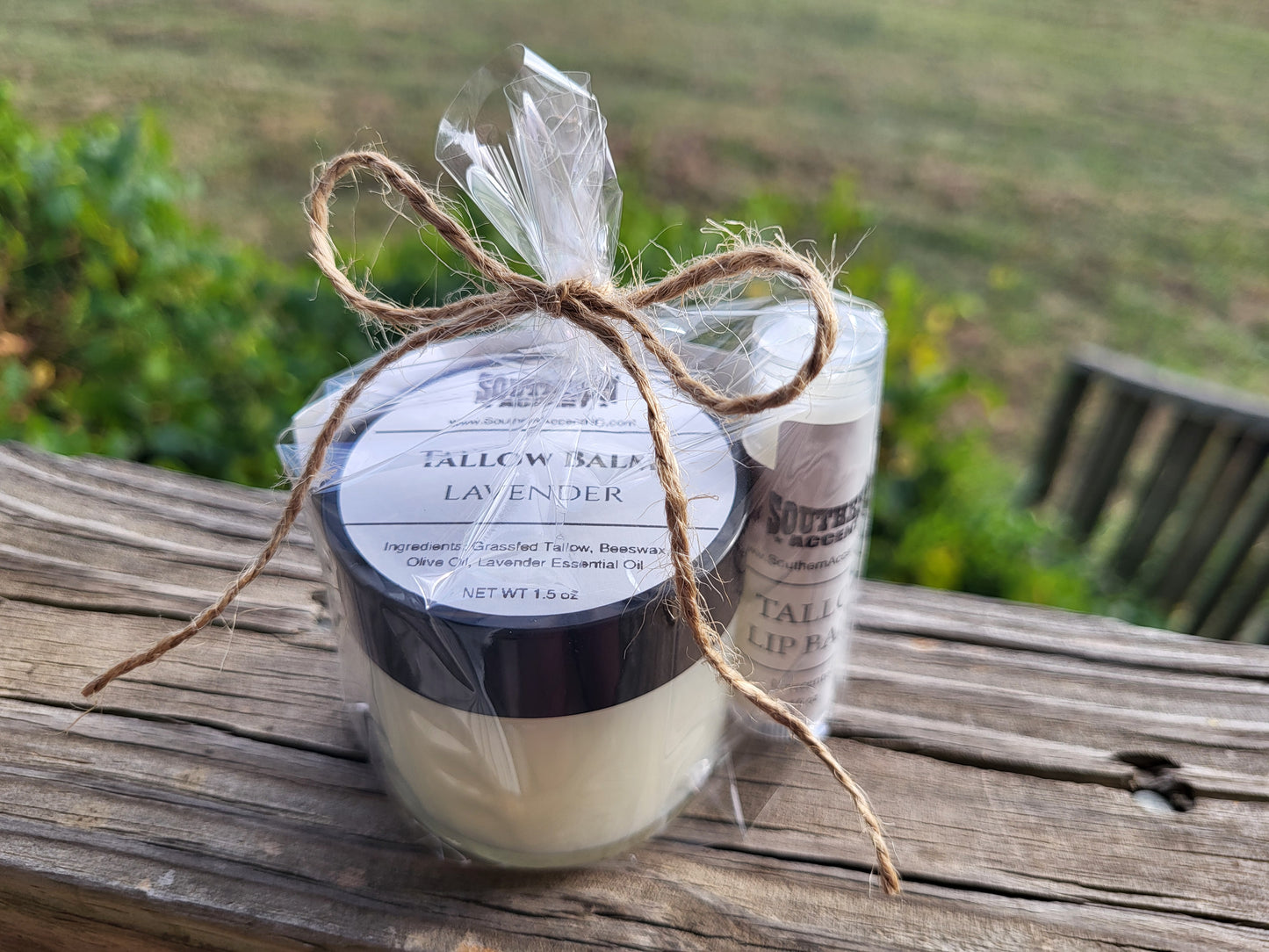 Clear jar of lavender Southern Accent brand Tallow Balm with a lip balm gift wrapped in a clear bag and tied with jute string sitting on a rustic wooden surface with a blurred natural background