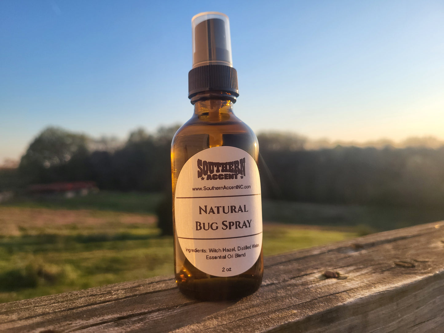 Southern Accent Natural Bug Spray 2oz amber glass bottle on a weathered wood surface with a sunny outdoor field in the background, natural DEET-free insect repellent