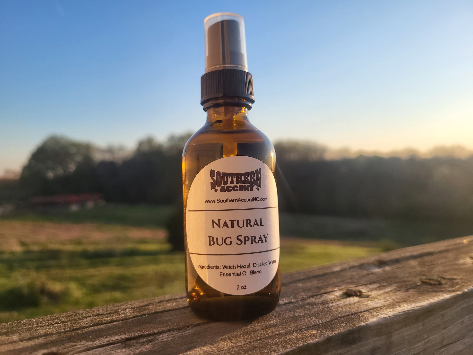 Southern Accent Natural Bug Spray 2oz amber glass bottle on a weathered wood surface with a sunny outdoor field in the background, natural DEET-free insect repellent
