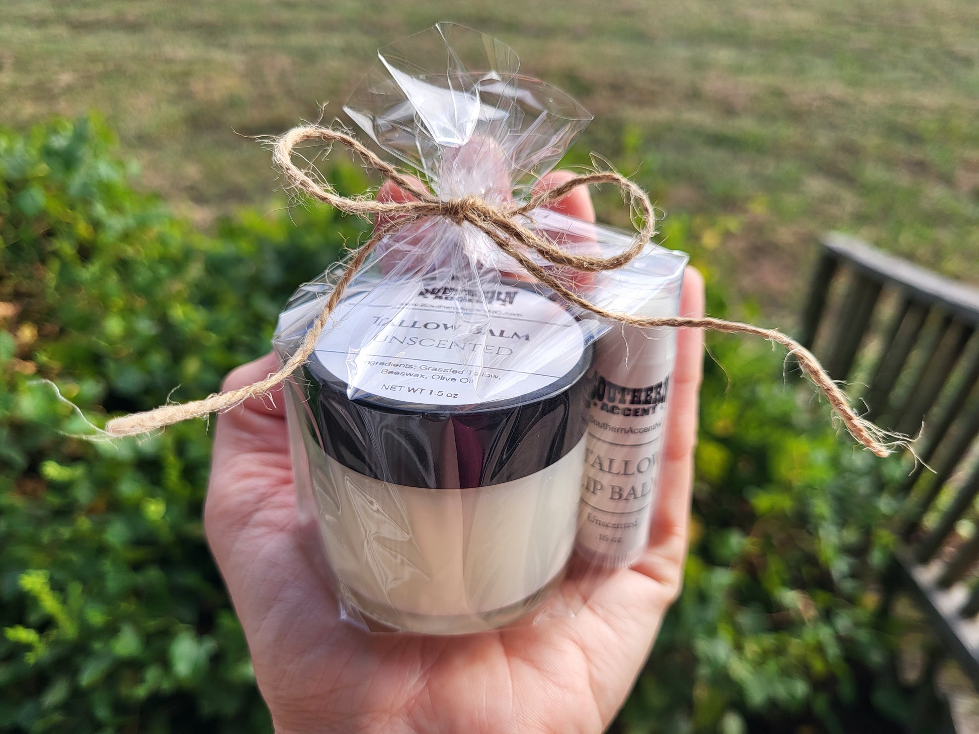 Hand holding a jar of southern accent tallow balm and lip balm wrapped in cellophane with twine, against a blurred natural background.